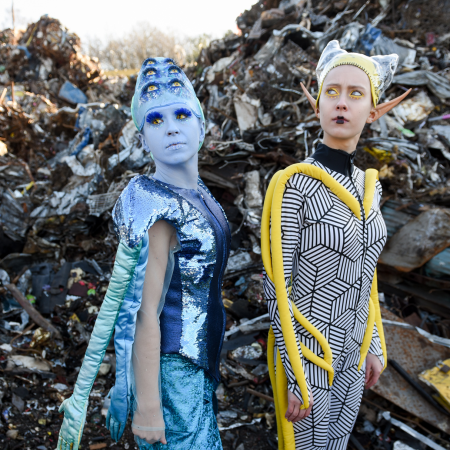 Dwie aktorki przebrane za kosmitki stoją na tle hałdy śmieci