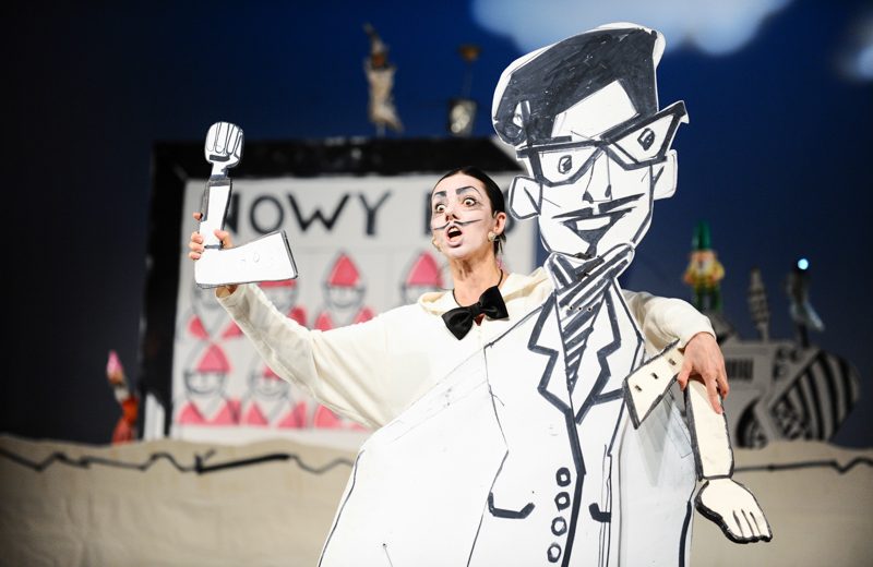 Aktorka ubrana na biało z czarną muszką i przyklejonymi wąsami stoi za dużą makietą rysunkowego mężczyzny w okularach
